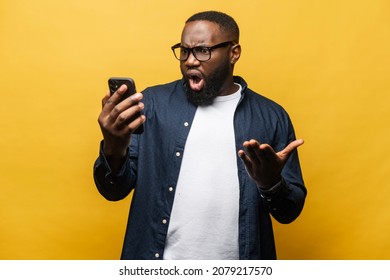 Shocked Surprised African-American Man Staring At Smartphone With Wide Open Mouth Isolated On Yellow Background. Black Guy Feels Negative Emotions, Amazed Facial Expression