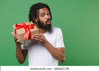 Shocked surprised curious young african american man with dreadlocks wearing white casual t-shirt posing hold red present box with gift ribbon bow isolated on green color background studio portrait - Powered by Shutterstock