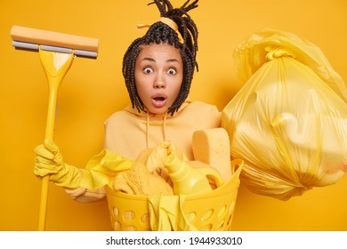 Shocked Surprised Ethnic Housemaid Wears Rubber Gloves For Hand Protection Holds Mop Trash Bag Basin Of Dirty Laundry Dressed In Hoodie Isolated Over Yellow Background. Cleaning Service Concept