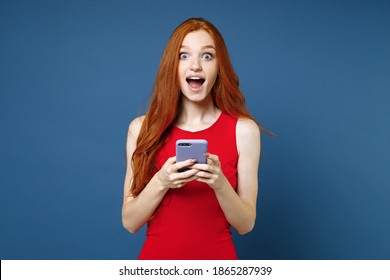 Shocked Surprised Excited Young Redhead Woman 20s Wearing Bright Red Elegant Evening Dress Standing Using Mobile Cell Phone Typing Sms Message Isolated On Blue Color Wall Background Studio Portrait