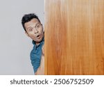 Shocked and surprised face of asian man peeking behind wooden cupboard.