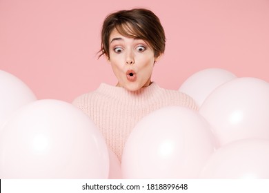Shocked surprised fun young brunette woman 20s in knitted casual sweater looking down hold bunch of air balloons, celebrating birthday holiday party say wow isolated on pastel pink background studio.
