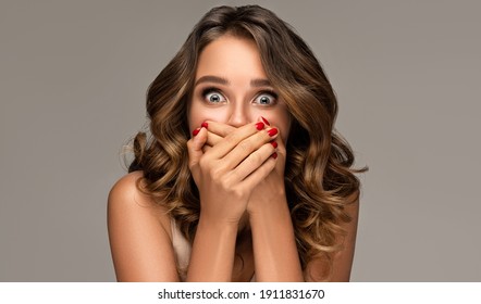 Shocked And Surprised Girl Screaming Covering  Mouth Her Hands.Curly Hair Woman Amazed.Beautiful Girl  With Wavy Hairstyle And Red Nails Manicure.Presenting Your Product.Expressive Facial Expressions