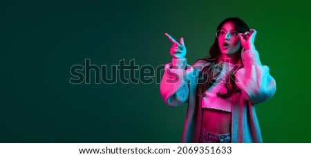 Shocked, surprised. One young adorable cute girl, student in casual style clothes posing isolated on green backgroud in pink neon light. Emotions, facial expression, youth, fashion and ad. Flyer