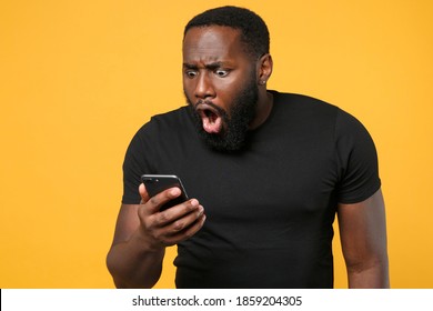 Shocked Surprised Upset African American Man In Casual Black T-shirt Isolated On Yellow Background Studio Portrait. People Lifestyle Concept. Mock Up Copy Space. Using Mobile Phone Typing Sms Message