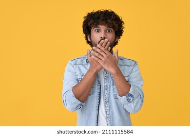 Shocked surprised young indian man looking at camera with omg face expression covering mouth with hands feeling amazed speachless silent reaction standing isolated on yellow background.
