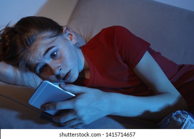 Shocked teenage girl with smartphone on sofa in dark room. Danger of internet