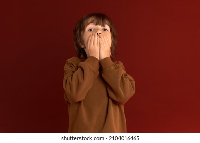 Shocked, Terrified And Scared Caucasian Boy In Brown Sweater Covering Mouth, Trying To Keep Silence, Be Quiet, Isolated On Red Background With Copy Space For Your Advertising Content