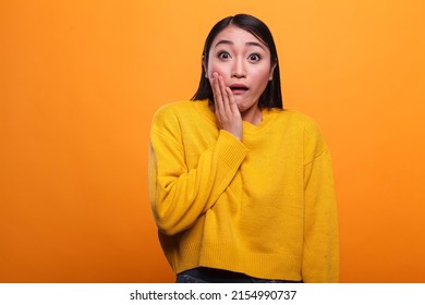Shocked troubled asian woman wearing yellow vibrant sweater while putting hand on face. Uneasy young adult girl with shock expression, wearing vivid trendy clothing while on orange background. - Shutterstock ID 2154990737