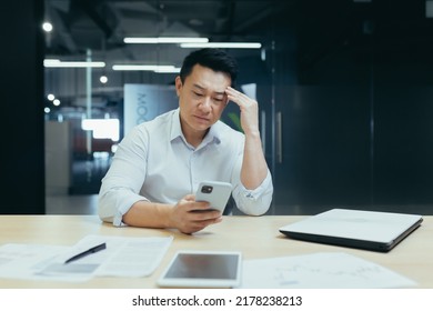 Shocked And Upset Young Man Asian Businessman Playing Games On Mobile Phone For Money At Work,lost,received Bad News By Message. Sitting Sad At The Desk In A Modern Office.