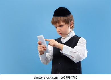 Shocked using phone. Portrait of a young orthodox jewish boy isolated on blue studio background. Purim, business, festival, holiday, childhood, celebration Pesach or Passover, judaism, religion