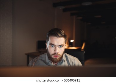 Shocked Web Designer Dressed In Shirt And Holding Eyeglasses Working Late At Night And Looking At Computer.