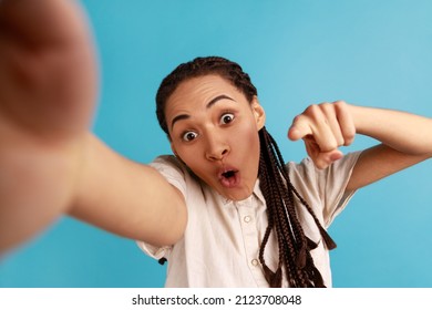 Shocked Woman With Black Dreadlocks Has Astonished Face Expression, Making Selfie, Expresses Surprised Emotions, Pointing Finger To Camera, POV. Indoor Studio Shot Isolated On Blue Background.