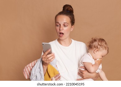 Shocked Woman With Bun Hairstyle Wearing White T Shirt Standing With Baby Kid And Using Cell Phone, Reading Surprised News, Looking At Screen With Big Eyes, Isolated Over Brown Background.