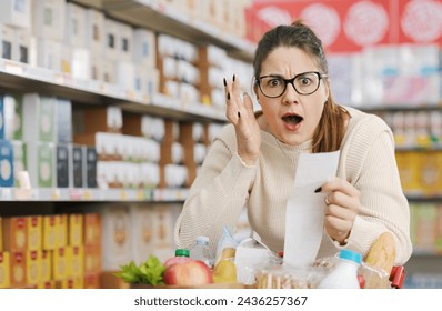 Shocked woman checking a long grocery receipt at the supermarket, expensive grocery and inflation concept - Powered by Shutterstock