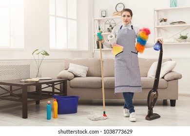 Shocked Woman Cleaning House With Lots Of Tools. Young Girl In Uniform Surprised Of Detergents Quantity And Mess In House, Copy Space