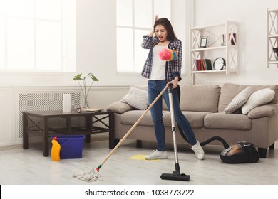 Shocked Woman Cleaning House With Lots Of Tools. Young Tired Girl Washing Floor With Mop And Vacuum Cleaner, Copy Space