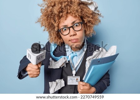 Shocked woman with curly hair stares at camera wears spectacles and formal outfit holds folders and microphone prepares for taking interview or giving speech during live stream. Mass media concept