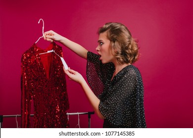 Shocked woman looking at price on sparkle red dress in boutique. Indoor portrait of amazed young female model holding hanger with expensive attire.