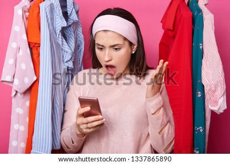 Shocked woman looks at smartphone display. Interesting information on screen. Dark haired lady raises palm and looks surprisingly at her gadget. Lots clothes behind beautiful female in shopping mall.