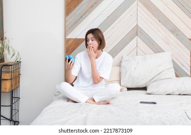 Shocked Woman Sitting On The Bed And Holding Alarm Clock. Waking Up Too Late Concept