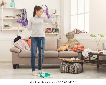 Shocked Woman With Special Equipment Cleaning House. Young Girl Surrounded By Many Stack Of Clothes. Disorder And Mess At Home, Copy Space