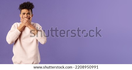 Shocked worried, embarrassed young man said something he shouldnt have, shut his mouth with hands and look guilty or anxious at camera, feel sorry for being rude, purple background