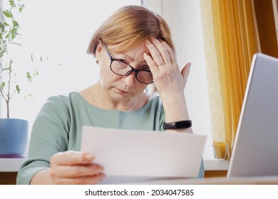 Shocked Worried Mature Woman Reading Paper Document Or Postal Letter Feeling Frustrated By Unexpected News Sitting At Home Indoor. Received Bank Account Balance Or Bad Notice, Fine, Bankruptcy