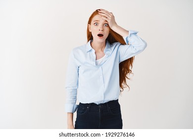 Shocked And Worried Redhead Woman Gasping And Staring At Camera Startled, Slap Forehead As Forget Something, Standing Over White Background Alarmed.