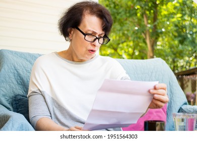 Shocked Worried Senior Woman Reading Paper Document Or Postal Letter Feeling Frustrated By Unexpected News Sitting On Sofa At Home Terrace.Received Bank Account Balance Or Bad Notice, Fine,bankruptcy