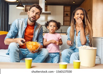 Shocked Young Adult Father And Mother Sitting On Comfort Couch With Cute African Child Girl, Watching Tv, Making Surprised Faces. Family Eating Chips And Spending Day Together At Home