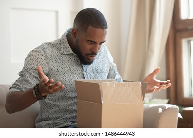 Shocked Young African American Male Customer Unpacking Delivered Cardboard Parcel With Order From Internet Online Store, Negatively Surprised Confused Angry Client Received Wrong Or Damaged Item.