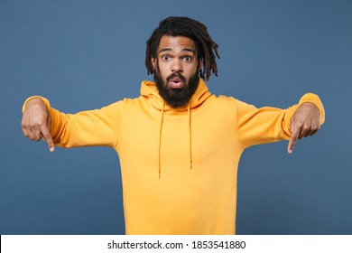 Shocked young african american man guy wearing yellow streetwear hoodie posing isolated on blue background studio portrait. People lifestyle concept. Mock up copy space. Pointing index fingers down