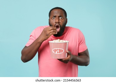 Shocked young african american man 20s in casual pink t-shirt isolated on blue background studio. People sincere emotions in cinema, lifestyle concept. Watching movie film, holding bucket of popcorn - Powered by Shutterstock