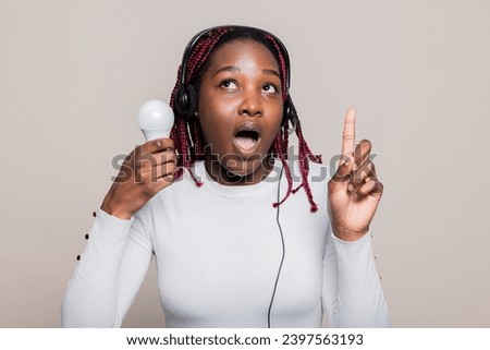 Shocked young African American woman with braids showing index finger at camera opening mouth having an idea holding bulb in hand creativity time.