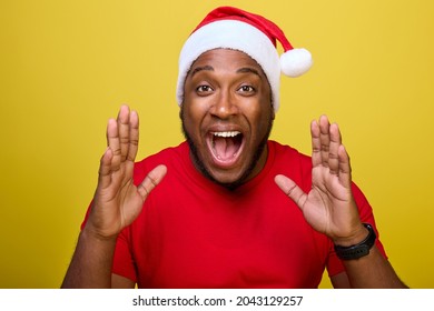 Shocked young African-American Santa Claus in a red T-shirt and a Christmas hat, shouting with a hand gesture at his mouth, isolated on a yellow background. Concept Happy New Year, Merry Christmas - Powered by Shutterstock