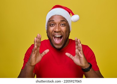 Shocked young African-American Santa Claus in a red T-shirt and a Christmas hat, shouting with a hand gesture at his mouth, isolated on a yellow background. Concept Happy New Year, Merry Christmas - Powered by Shutterstock