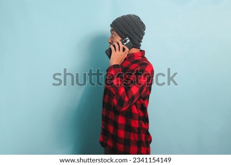 Shocked Young Asian man with beanie hat and red plaid flannel shirt is surprised, with his hand on his waist in an arm akimbo position, while talking on his mobile phone, isolated on a blue background