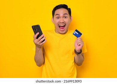 Shocked Young Asian Man In Casual T-shirt Holding Mobile Phone And Credit Card Isolated On Yellow Background. People Lifestyle Concept