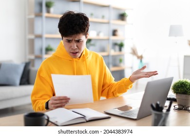 Shocked Young Asian Man Sitting At Table With Modern Laptop At Home Office, Reading Paper Letter Mail And Spreading Hands, Holding Document Or Bills, Pensive Guy Got Bad News, Debt Notification