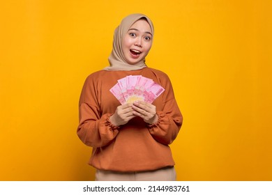 Shocked Young Asian Woman In Orange Shirt Holding Money Banknotes Isolated Over Yellow Background