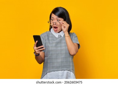 Shocked young Asian woman using mobile phone and taking of glasses isolated over yellow background