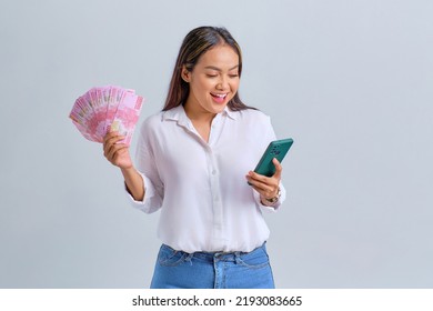 Shocked Young Asian Woman Using Mobile Phone And Holding Money Banknotes Isolated Over White Background