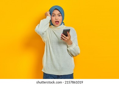 Shocked Young Asian Woman In White Sweater And Hijab Using On Mobile Phone And Holding Head With Hand Isolated Over Yellow Background