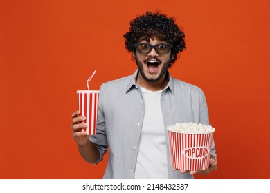 Shocked young bearded Indian man 20s wears blue shirt keeping mouth wide open in 3d glasses watch movie film hold bucket of popcorn cup of soda pop isolated on plain orange background studio portrait