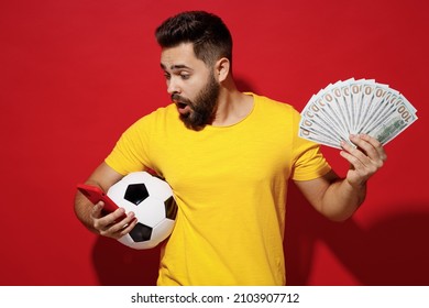 Shocked Young Bearded Man Football Fan In Yellow T-shirt Cheer Up Support Favorite Team Hold Soccer Ball Fan Of Cash Money Look At Mobile Cell Phone Isolated Plain Dark Red Background Studio Portrait