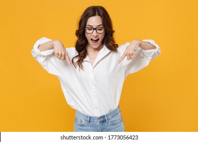 Shocked young brunette business woman in white shirt glasses isolated on yellow background studio portrait. Achievement career wealth business concept. Mock up copy space. Pointing index fingers down