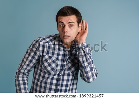 Shocked young brunette guy looking aside and overhearing isolated over blue background