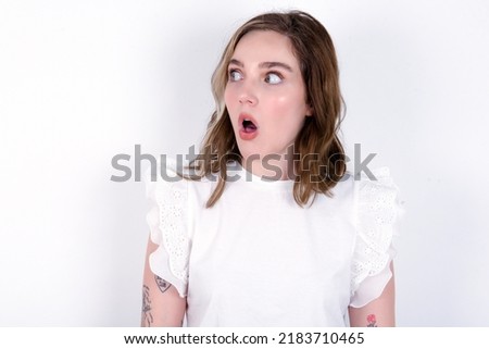 Shocked young caucasian woman wearing white T-shirt over white background look empty space with open mouth screaming: Oh My God! I can't believe this.