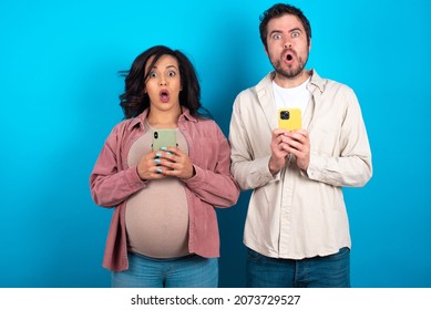 Shocked Young Couple Expecting A Baby Standing Against Blue Background Opens Mouth Hold Phone Reading Advert Unbelievable Big Shopping Prices